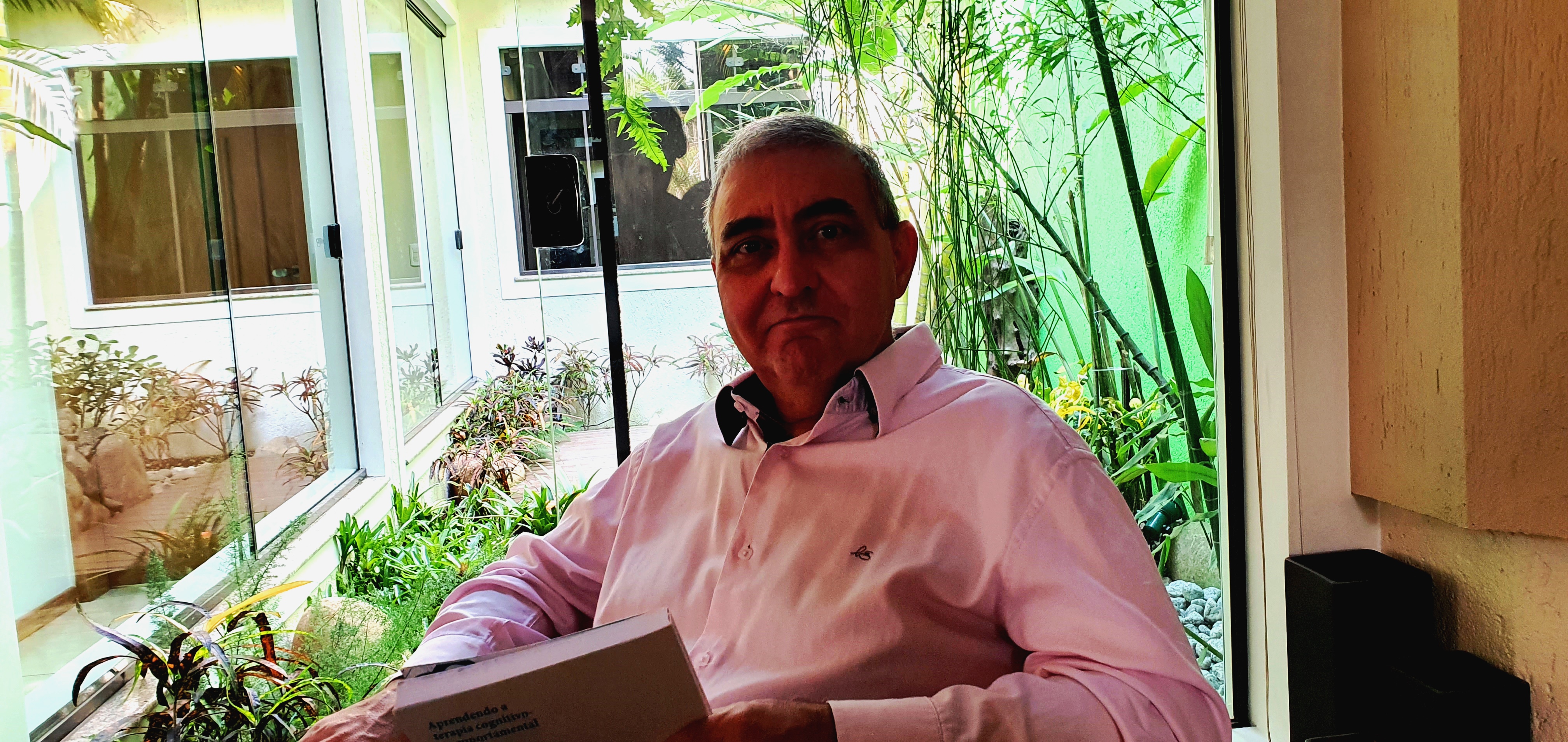Man sitting in front of a window in a white brick or plaster building, pastel pink shirt, book in front of him. Here's the...